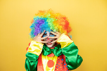 Clown standing over yellow insolated yellow background peeking in shock covering face and eyes with hand, looking through fingers with embarrassed expression - obrazy, fototapety, plakaty