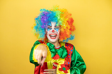 Clown standing over yellow insolated yellow background laughing at you, pointing finger to the...