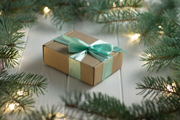 Fototapeta na wymiar Gift wrapped in craft paper and mint ribbon with green branches of Christmas tree with bulbs on white wooden background.