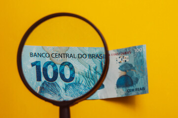 Brazilian money being viewed through a magnifying glass (in Brazil: LUPA), with notepad and calculator. Finance and salary concept. Economy of Brazil. Yellow background