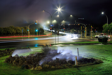 A main road in Rotorua, New Zealand, at night. Streetlights and car headlights light up steam from geothermal hot pools