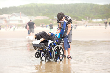 Obraz premium Young man pushing disabled little boy in wheelchair on beach