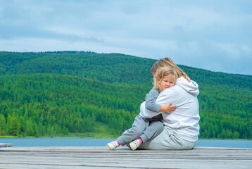 Mom hugs a child sitting on the pier of a blue lake in the mountains. Family tourism