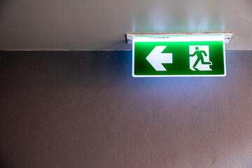 Fire exit sign with icon and arrow pointing to the left on green background hanging from the ceiling.
Exit sign on a wall.