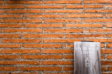"Entrance Only" sign on wooden plank in English and Japanese texts leaning against orange brick wall.
