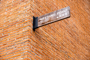 "Meeting Point" sign on wooden plank in English and Chinese languages attached to orange brick wall.
