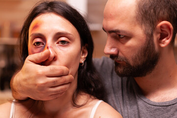 Violent husband covering wife mouth after beating and bruising her face. Aggressive husband abusing injuring terrified helpless, vulnerable, afraid, beaten and panicked woman.