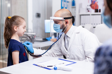 Pediatrician wearing face mask agasint coronavirus listening child heart with stethoscope during consultation. Health doctor specialist providing health care services consultations.