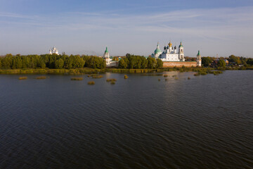 Aerial view of a Russian Orthodox monastery on a lakeside surrounded by grass and trees in autumn colours. Light white clouds on blue sky. No people.