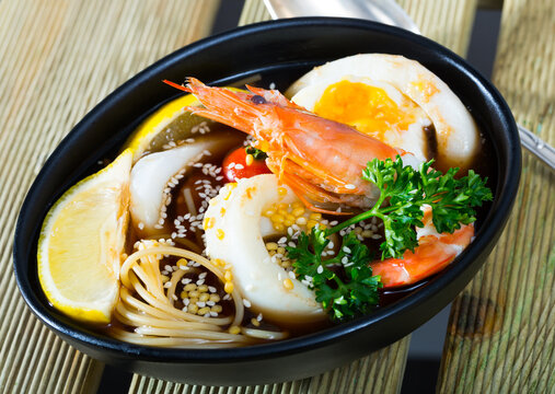 Photography of plate with spicy pan-Asian soup with squid, shrimp, egg noodles and sesame in restaurante.