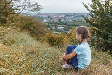 Naklejka na ściany i meble Little girl sitting on a hill above the city. The child sits embracing her legs in the tall grass