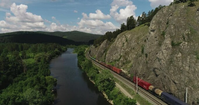 Freight long train carries with cargo carriages under the rocky mount in wild mountains landscape through a difficult part of Trans Siberian railways. Aerial drone wide view at summer day.