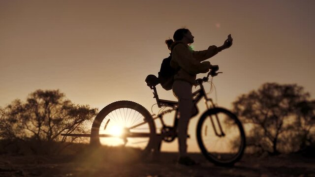 teenage girl photographs herself on a smartphone with a bicycle rides in the park at sunset on nature. healthy girls and bike makes selfie lifestyle