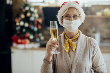 Joyful senior woman with face mask raising a glass while celebrating New Year at home.