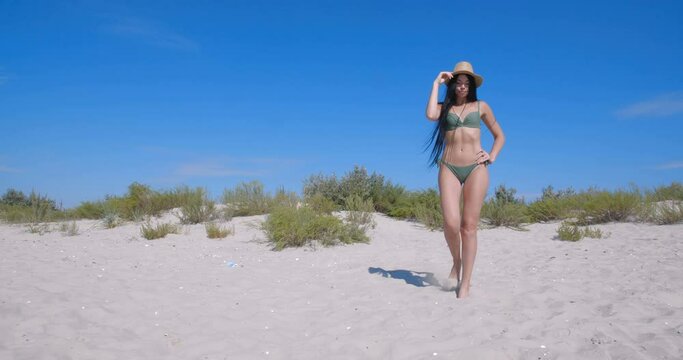 Attractive tall brunette in green swimsuit and straw hat walking on a beach. Beautiful mixed race model with very long black hair posing on a beach. Full-height shooting. 4k 50fps slow motion