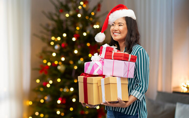 Obraz na płótnie Canvas winter holidays and people concept - happy asian young woman in santa helper's hat with gift boxes over christmas tree at home on background