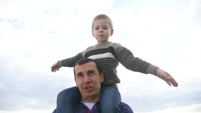 happy family. son sits on his neck teamwork at father shows hands to the side plays at the pilot depicts an airplane. happy family concept childhood man lifestyle dad with little boy son walk on field