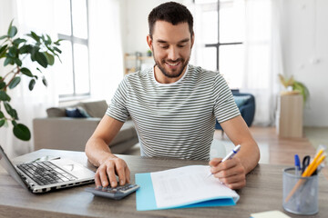 remote job and business concept - happy smiling man with papers, calculator and laptop computer working at home office