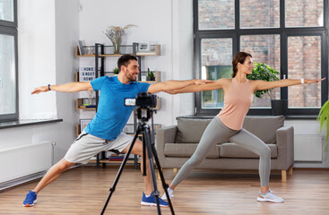 fitness, sport and video blogging concept - couple of bloggers with camera on tripod recording online yoga class at home