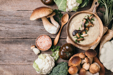 Wooden bowl of cream mushroom soup with fried mushrooms, vegetables, spices, raw boletus edulis mushroom on wooden background. Top view. Copy space. Autumn harvest concept
