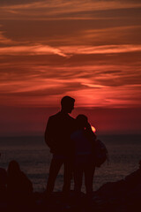 Fototapeta na wymiar Young couple in love admiring a beautiful red and orange sunset in a famous sunset point in Sardinia, Italy. Romantic moment for the two, framed by the sun an the scenic sky.