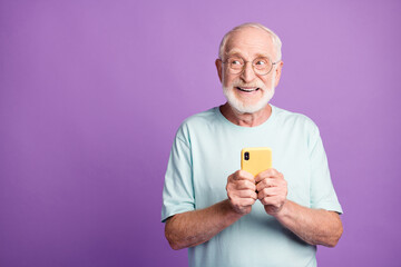 Photo portrait of elderly man holding phone in two hands looking at blank space isolated on vivid purple colored background