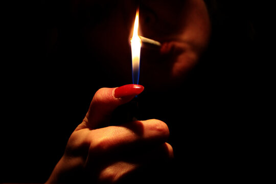 Part of woman face with cigarette, fingers with red nails lightning the cigarette in darkness.