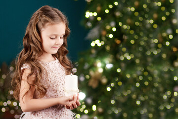 Portrait of acute long-haired little girl in dress on background of  lights.Little girl holding burning candle. Christmas, New Year and birthday celebration concept. Winter holidays. Copy space