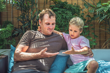 Father blond son sit look on open hands, play compare their palms. Child gently hugs daddy by neck. Bright sun beams light leaves garden terrace. Happy childhood, fatherhood love feelings, fathers day