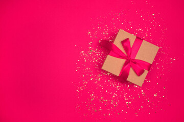 Gift box with red bow on festive bright pink background with golden sparkles around and copyspace for your text. Flat lay style. Christmas, New Year, Valentines Day or birthday celebration concept