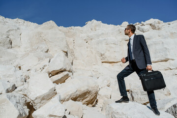 Young man businessman with a briefcase and jacket is climbing the mountain.