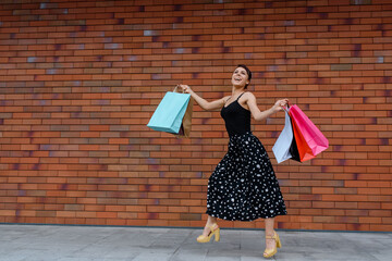 Young happy smiling beautiful woman is jumping and running on the street near a brick wall and holding a lot of shopping bags.