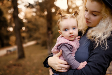Fototapeta premium Young mother is holding her 6 months old baby girl in her arms in the autumn park.