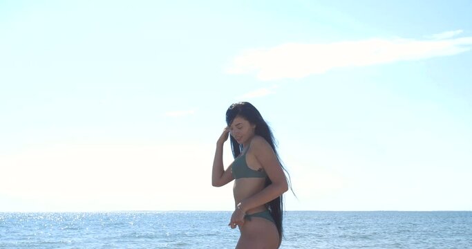 Attractive tall brunette in green swimsuit posing on the seashore flirting to camera. Rear view of beautiful mixed race model with very long black hair  on a beach.  4K 50fps slow motion