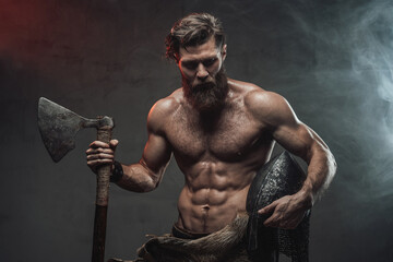 Serious and muscular viking with beard and naked torso staying in dark background with lights and...