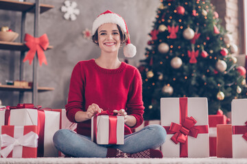 Obraz na płótnie Canvas Photo of lovely girl sit carpet receive giftbox toothy smile look camera wear santa cap red pullover decorated living room indoors