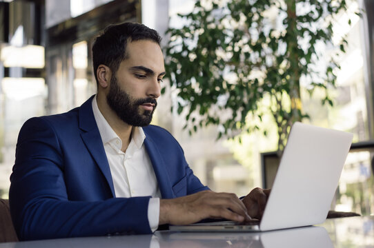 Young handsome arabic businessman planning project, typing on keyboard in modern office. Portrait of pensive middle eastern man wearing formal clothes, working online, sitting at workplace