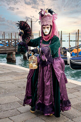 Obraz premium Woman in mask wearing ornate colorful carnival costume on San Marco lagoon background at sunset. Carnival in Venice, Italy