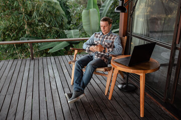 Young caucasian man outside a cafe looking at his smartphone, sit by the table with laptop in cafe terace with nature view