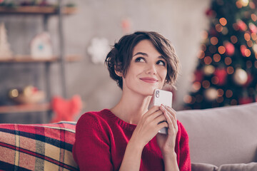 Photo of inspired minded girl sit cozy couch hold smartphone think look in house indoors with x-mas...