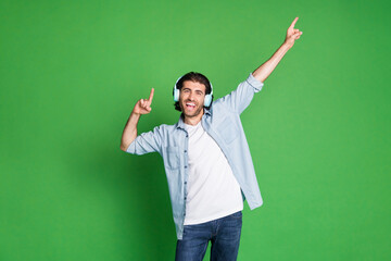 Photo of stubbled young guy raise hands indicate empty space wear headphones denim shirt jeans isolated green color background