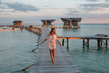 Back view young woman walking on a wooden pier to the bungalow in the ocean. Attractive woman walks on a wooden jetty towards a tropical island during sunset