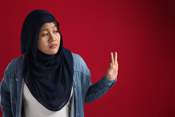 Asian muslim woman wearing hijab rejecting avoiding something from the side, red background with...