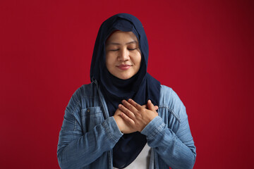Calm Asian muslim female with closed eyes, hands on heart, feels gratitude, happy smiling expression