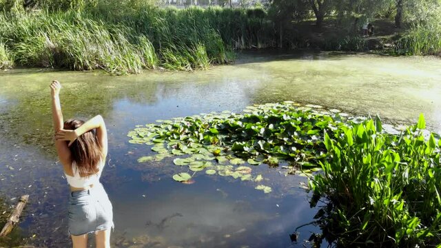Rear view of a sexy girl standing near green pond and forest. Clip. Attractive young woman in white top and denim mini skirt dancing slowly on the shore of a mossy pond water.