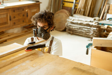 A young man working as carpenter in his wood workshop. Wood worker designing and handcrafting new house furniture using a piece of timber and wearing a full face mask for the dust