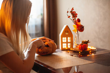 Woman carves pumpkin for Halloween in room with autumn decor and lamp house. Cosy home and preparing for Halloween.