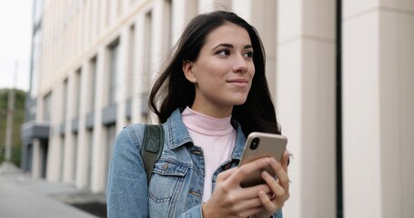 Close up portrait of cheerful young Caucasian female standing in city and tapping on smartphone. Beautiful alone joyful woman typing and browsing on cellphone outdoors and smiling. Leisure concept