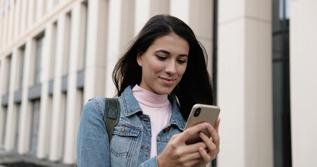 Close up portrait of cheerful young Caucasian female standing in city and tapping on smartphone. Beautiful alone joyful woman typing and browsing on cellphone outdoors and smiling. Leisure concept