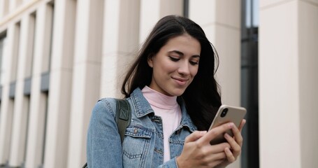 Close up portrait of cheerful young Caucasian female standing in city and tapping on smartphone. Beautiful alone joyful woman typing and browsing on cellphone outdoors and smiling. Leisure concept